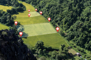 Construction Layout Surveys leads to Aerial photo of green rice fields divided with dotted boundary lines and red location pins, surrounded by tropical forest and mountains in rural farming area, suitable for agricultural planning.